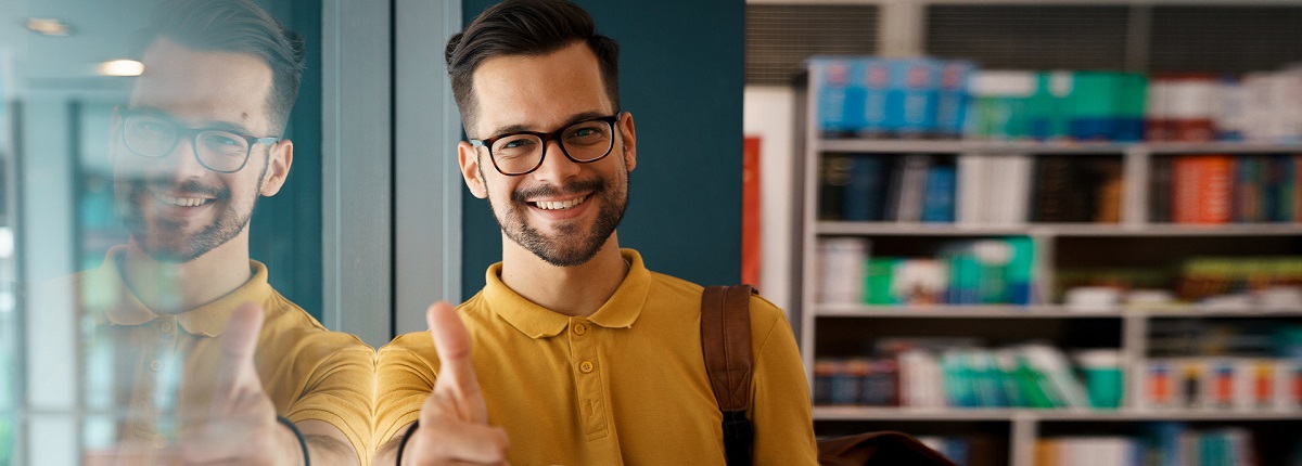 <p>Ein junger Mann mit senfgelbem Hemd steht in einem Büro und lehnt sich an eine Glaswand. Er schaut freundlich lächelnd in die Kamera. Im Hintergrund ist ein Regal zu erkennen.</p>